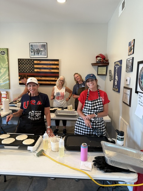 Five women smile while cooking pancakes on griddles in a room decorated with American flags and firefighter memorabilia. Tables hold cooking supplies, and the women wear casual clothes and aprons.