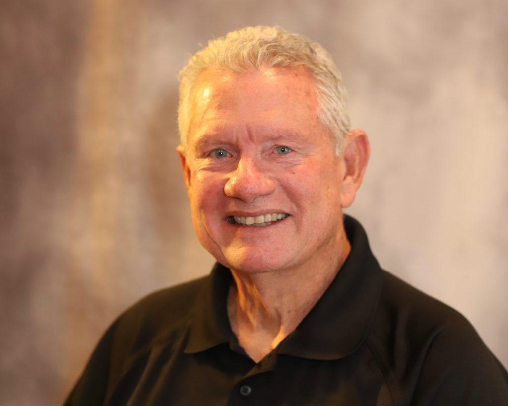 An older man with short gray hair smiles at the camera. He is wearing a black collared shirt and is posed in front of a blurred, neutral-colored background.
