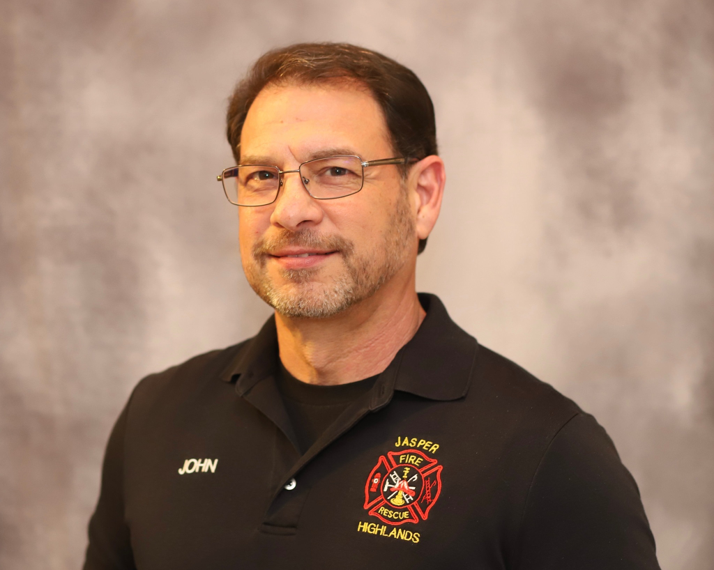 A man with short brown hair, glasses, and a trimmed beard wears a black polo shirt embroidered with "Jasper Highlands Fire Rescue" and the name "John." He stands in front of a gray, softly blurred background.