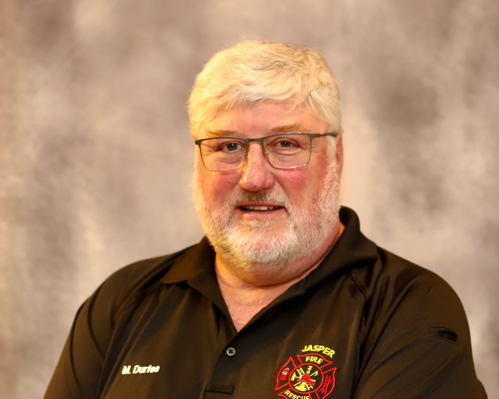A man with white hair and a beard, wearing glasses and a black Jasper Fire Department polo shirt, smiles in front of a softly blurred gray background.