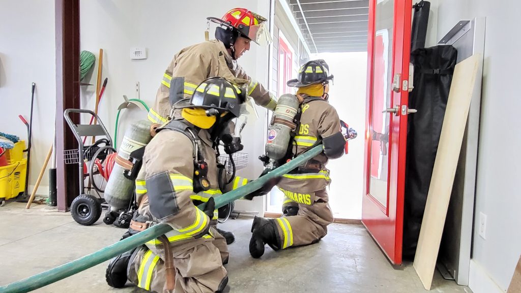 Three firefighters in full gear kneel indoors, holding a fire hose and preparing to head outside through an open door. Equipment and cleaning supplies are visible in the background.