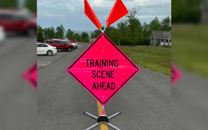 A bright pink diamond-shaped road sign with two orange flags reads "TRAINING SCENE AHEAD" and is positioned on a road, with parked cars, trees, and a building in the background.