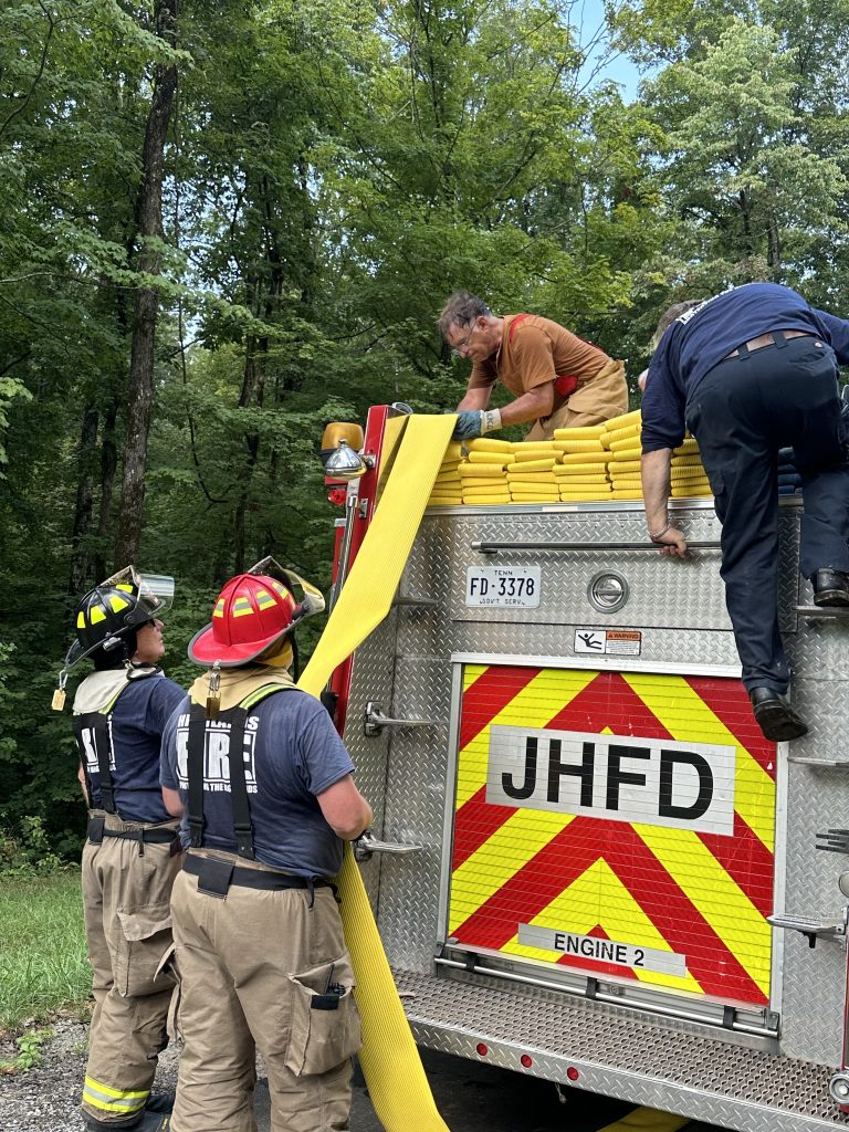Three firefighters in gear stand by a fire truck while two people on the truck work with yellow fire hoses. The scene is outdoors, surrounded by green trees. The truck displays “JHFD” and “Engine 2” on the back.